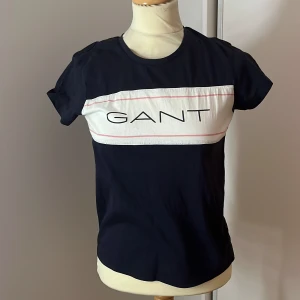 Mörkblå och vit t-shirt från GANT - Snygg mörkblå t-shirt från GANT med vit panel och logga över bröstet. Den är kortärmad och har en klassisk rund halsringning. Perfekt för en avslappnad stil eller vardagsbruk. Passar bra till jeans eller shorts!