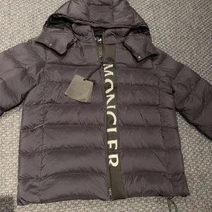 Svart dunjacka från Moncler - Säljer en snygg svart dunjacka från Moncler i mycket bra skick. Jackan har en avtagbar huva och dragkedja framtill. Perfekt för kalla vinterdagar med sin varma och puffiga design. Moncler-loggan syns tydligt på ärmen. Passar både till vardags och lite finare tillfällen.