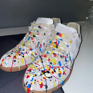 Maison Margiela skor - Säljer ett par unika Maison Margiela GATs i vitt med ett coolt stänkmönster i rött, blått, gult och svart. Skorna har en klassisk design med snörning och en bekväm brun sula. Perfekta för att ge din outfit en färgklick! De är i helt nytt skick! Skriv järna om du är intresserad!