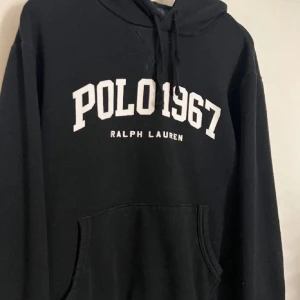 Svart hoodie från Ralph Lauren - Säljer en klassisk svart hoodie från Ralph Lauren med texten 'POLO 1967' i vitt på framsidan. Den har en bekväm passform och en praktisk magficka. Perfekt för en avslappnad stil eller kyliga dagar. Huvan har justerbara snören för extra komfort.