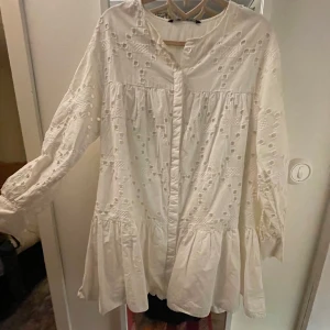 Vit klänning med broderade detaljer - Säljer en superfin vit klänning med broderade detaljer och volanger. Den har långa ärmar och en lös passform, perfekt för sommar och student. Klänningen är i mycket bra skick. Perfekt för både vardag och festliga tillfällen!