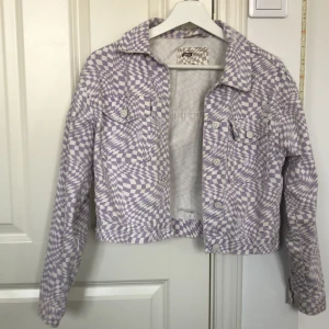 Lila och vit rutig jacka från New Yorker - Säljer en cool lila och vit rutig jeansjacka från New Yorker i storlek XS. Jackan har en croppad stil med knappar framtill och på ärmsluten. Den har lite håll överallt som är en del av designen. Perfekt för vår och sommar!