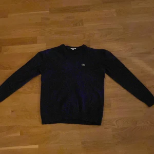 Marinblå tröja från Lacoste - Säljer en stilren marinblå tröja från Lacoste i mycket bra skick. Den har en klassisk v-ringning och det ikoniska krokodilmärket på bröstet. Perfekt för både vardag och lite finare tillfällen. Passar bra till höst och vår.