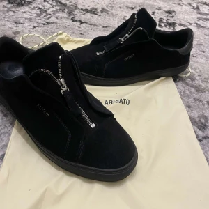 Svarta sneakers från Axel Arigato - Snygga svarta sneakers från Axel Arigato i mocka med dragkedja istället för snörning. Perfekta för en stilren look, oavsett om det är vardag eller fest. De är i mycket bra skick och kommer med originalpåse.