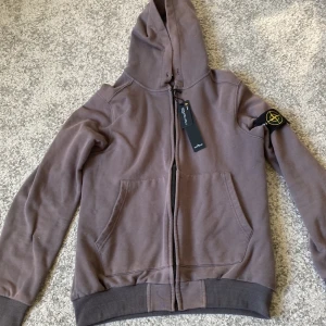 Brun hoodie från Stone Island - Säljer en snygg grå hoodie från Stone Island. Den har en dragkedja framtill och en klassisk huva. På ärmen finns det ikoniska Stone Island-märket. Perfekt för höst och vår! snöre med följer också. M passar S bäst