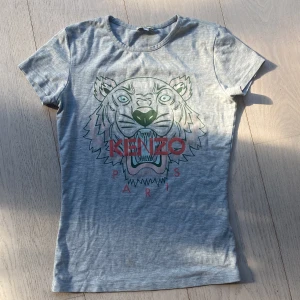 Grå t-shirt från Kenzo - Säljer en snygg grå t-shirt från Kenzo med ett coolt tigertryck på framsidan. T-shirten är i storlek 12A/152 och har korta ärmar. Perfekt för en avslappnad stil! 