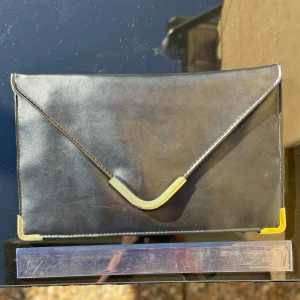 Svart kuvertväska med gulddetaljer - Svart kuvertväska i syntet med snygga gulddetaljer på hörnen och en stilren design. Väskan har en knappstängning. Den är i bra skick och redo att användas! Mått se linjalen på bilderna. Hämtas på Hägerstensåsen eller ök 🖤