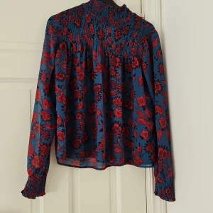 Blommig blus i blått och rött - Säljer en superfin blommig blus i blått och rött. Den har långa ärmar och ett smockat parti upptill som ger en snygg passform. Perfekt för både vardag och fest! 🌸