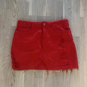 Röd jeanskjol med slitningar - Säljer en cool röd jeanskjol med slitningar. Kjolen är kort och har en rå kant nertill som ger en edgy look. Den har fickor både fram och bak, samt en knapp och dragkedja framtill.