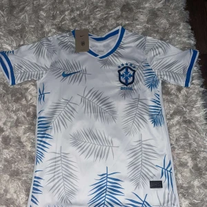 Vit och blå fotbollströja från Nike - Säljer en snygg vit fotbollströja från Nike med blå detaljer. Tröjan har ett coolt mönster av palmblad i grått och blått, och det brasilianska fotbollsförbundets emblem på bröstet. Den är kortärmad och perfekt för både träning och match. Helt ny i förpackning!