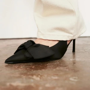 Svarta klackskor med rosett från Zara - Snygga svarta klackskor från Zara med spetsig tå och en elegant rosett på framsidan. Skorna har en smal klack och en stilren design som passar perfekt för festliga tillfällen. De är tillverkade i ett syntetmaterial som ger en lyxig känsla. Aldrig använda! 