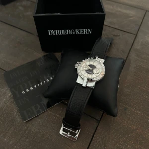 Elegant klocka från Dyrberg/Kern - Säljer en superfin klocka från Dyrberg/Kern. Den har en stilren design med en svart läderrem och ett silverfärgat urverk. Urtavlan är dekorerad med glittriga stenar runt kanten och har en unik design med två mindre urtavlor inuti. Perfekt för både vardag och fest! Kommer med originalförpackning och certifikat.