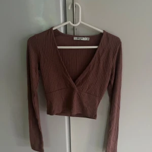 Brun ribbad topp från Na-Kd - Säljer en snygg brun ribbad topp från Na-Kd. Den har en djup v-ringning och är croppad, vilket gör den perfekt för både vardag och fest. Toppen har långa ärmar och är i ett mjukt och stretchigt material.