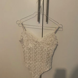 Vit spetsbody - Säljer en superfin vit spetsbody med blommigt mönster. Den är ärmlös och har smala axelband, perfekt för fest eller sommarens alla tillfällen. Bodyn är i storlek M och sitter tight för en snygg passform.