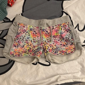 Grå vintage shorts med färgglatt mönster - Säljer ett par grå vintage shorts med färgglatt mönster på framsidan. De har en bekväm passform med elastisk midja och snörning. Perfekta för en avslappnad stil. De har ingen tagg men jag gissar storleken är XS/S