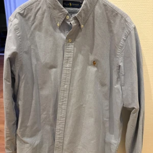 Grå skjorta från Ralph Lauren - Snygg grå skjorta från Ralph Lauren med klassisk button-down krage och broderad logotyp på bröstet. Skjortan har långa ärmar och knappar framtill. Perfekt för en stilren look.