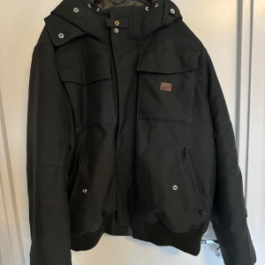 G Star Vinterjacka - Snygg svart bomberjacka med huva och flera fickor. Jackan har dragkedja och knappar framtill samt ribbade muddar vid ärmslut och nederkant. Perfekt för kyligare dagar.