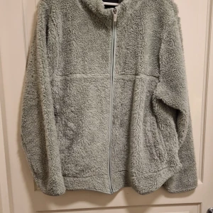 Grå teddyjacka från 157 - Mysig grå teddyjacka från 157 med dragkedja framtill. Perfekt för kyliga dagar med sin fluffiga och varma design. Jackan har långa ärmar och en avslappnad passform.