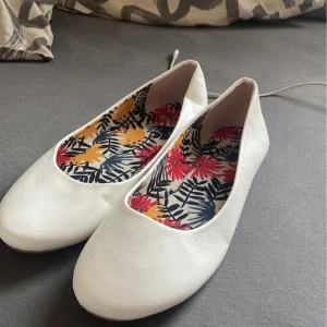 Vita ballerinaskor med blommigt mönster - Säljer ett par vita ballerinaskor med en färgglad blommig insida. Perfekta för en somrig look med sin enkla och stilrena design.
