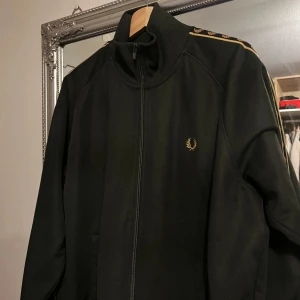 Svart jacka från Fred Perry - Säljer en snygg svart jacka från Fred Perry i mycket bra skick. Den har en dragkedja framtill och coola detaljer med Fred Perry-loggan längs ärmarna. Perfekt för höst och vår, och den har en normal passform. Jackan är långärmad och gjord i ett bekvämt material.