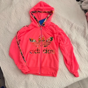 Rosa hoodie från Adidas Originals - Säljer en färgglad rosa hoodie från Adidas Originals med blommigt mönster på loggan och insidan av huvan. Den har dragkedja framtill och långa ärmar. Den är i storlek S