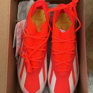 Adidas X Crazyfast.1 FG Fotbollsskor - Snygga och sportiga Adidas X Crazyfast.1 FG fotbollsskor i en iögonfallande röd färg med vita detaljer. Skorna har snörning och är designade för att ge optimalt grepp på fasta underlag med sina metalliska dobbar.