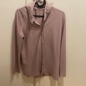 Rosa ribbad skjorta - Säljer en stilren rosa ribbad skjorta med knappar framtill. Skjortan har långa ärmar och en klassisk krage, perfekt för en chic look.