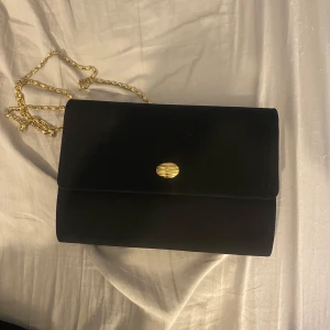 Svart kuvertväska med guldkedja från Vezzano - Elegant svart kuvertväska från Vezzano med en stilren guldkedja som axelrem. Väskan har en enkel knappstängning i guld och flera fack inuti för smidig organisering. Perfekt för en kväll ute eller en speciell tillställning.