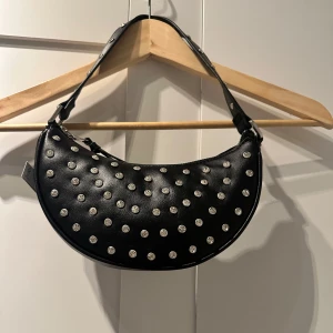 Svart axelväska med diamanter  - Snygg svart axelväska i syntetmaterial med diamanter. Väskan har en dragkedja upptill och en justerbar axelrem. Perfekt för att ge din outfit en edgy touch. Helt ny prislappen är fortfarande på den. Varumärket är Rita Ora X Primark. Jätte fin och bra skin väska. Perfekt för och cluba eller till någon fixad outfit.