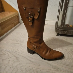 Bruna stövlar från Novita - Snygga bruna stövlar i skinn från Novita med dragkedja på sidan och dekorativt spänne upptill. Perfekta för höst och vinter med en klassisk design som passar till många outfits.