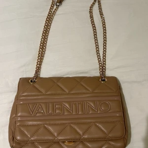 Beige quiltad axelväska från Mario Valentino - Snygg beige axelväska från Mario Valentino med quiltad design och kedjeaxelrem. Väskan har en elegant look med logotypen framtill och är perfekt för att lyfta din stil.