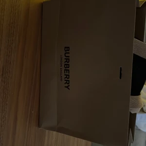 Svart keps från Burberry - Snygg svart keps från Burberry med vit broderad logotyp på framsidan. Skärmen har det klassiska Burberry-rutiga mönstret på undersidan. Perfekt för en stilren look.