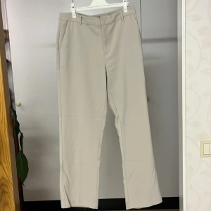 Beige kostymbyxor från Gina Tricot - Snygga beige kostymbyxor från Gina Tricot med en klassisk rak passform. Byxorna har en stilren design med hällor för bälte och en diskret dragkedja. Perfekta för en elegant look.