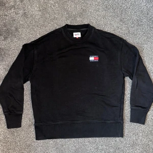 Svart sweatshirt från Tommy Jeans - Säljer en stilren svart sweatshirt från Tommy Jeans med deras ikoniska logga på bröstet. Tröjan har långa ärmar och en rund halsringning. Perfekt för en avslappnad look.
