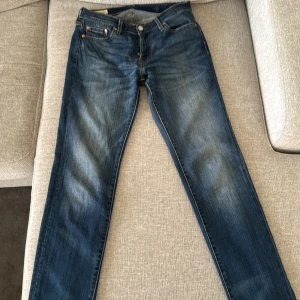 Blå jeans från Levi's - Säljer ett par klassiska blå jeans från Levi's med fem fickor och orange sömmar. De har en knappgylf och en läderpatch med logotyp på baksidan. Perfekta för en avslappnad stil.