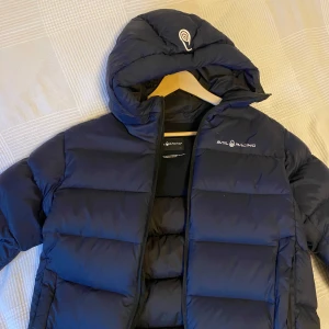 Mörkblå vinterjacka från Sail Racing - Snygg och varm mörkblå jacka från Sail Racing. Jackan har en dragkedja framtill och är perfekt för kyliga dagar. Den är långärmad och har en stilren design med logotyp på bröstet. Är i mycket fint skick, storlek 176 så passar typ S.