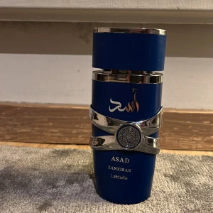 Asad Zanzibar parfym från Lattafa - Elegant parfymflaska i djupblått med silverdetaljer och arabiska inskriptioner. Flaskan har en unik form och ett dekorativt mönster på framsidan. Perfekt för den som söker en exotisk doftupplevelse.