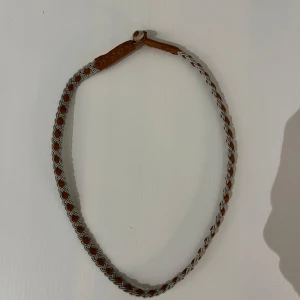 Flätat tenn halsband i läder och silver - Snyggt flätat tenn halsband med en kombination av brunt läder och silverfärgad metall. Perfekt för att ge en stilren touch till din outfit. Halsbandet har en enkel knäppning och passar de flesta tillfällen.