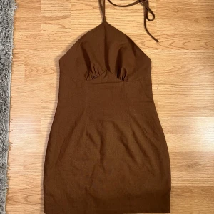 Brun klänning från NA-KD - Snygg brun klänning från NA-KD med halterneck-design. Perfekt för en stilren look. Klänningen har en åtsittande passform och är tillverkad i ett mjukt material. Rekommenderar till folk som är under 170 då det sitter ganska kort på mig personligen 