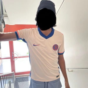 Chelsea fotbollströja  - Snygg vit fotbollströja från Nike med blå krage och röda detaljer. Tröjan har korta ärmar med blå och röda ränder. Perfekt för fotbollsfans som vill visa sitt stöd. 10/10 