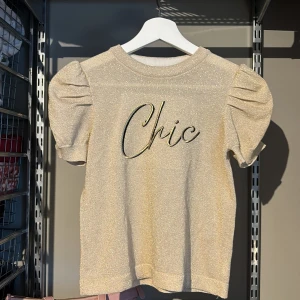 Glittrig topp från River Island.  - Snygg glittrig topp från River Island med texten 'Chic' på framsidan. Toppen har korta puffärmar och en rund halsringning. Perfekt för att lysa upp vilken outfit som helst! Även mycket bra kvalite, då den inte är använd! Om du köper både denna tröja och min kjol som ska säljas får du både för bara 100kr!