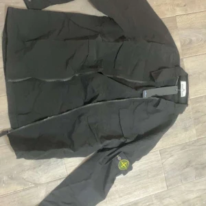 Svart windbreaker från Stone Island - Snygg svart jacka från Stone Island med dragkedja och broderad logga på ärmen. Perfekt för en stilren look. Skriv för mer info och bilder!