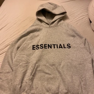 Grå Essentials hoodie från Fear of God - Säljer en grå Essentials hoodie från Fear of God. Den har en klassisk design med en stor logotyp på framsidan och en bekväm huva. Perfekt för en avslappnad stil.