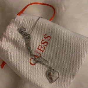 Silverfärgat armband från Guess - Elegant silverfärgat armband från Guess med ett hjärtformat hänge. Hänget har en glittrig kant och Guess-loggan ingraverad. Aldrig använd