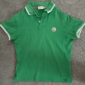 Grön pikétröja från Moncler - Snygg grön pikétröja från Moncler med vit randig krage och ärmslut. Tröjan har en klassisk pikédesign med knappslå och Moncler-logga på bröstet. Perfekt för en stilren look.