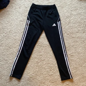 Svarta träningsbyxor från Adidas - Säljer ett par svarta träningsbyxor från Adidas med klassiska vita ränder längs sidorna. Byxorna har en elastisk midja och är perfekta för sport och aktivitet. De är tillverkade i ett lätt och bekvämt material. Storlek 152💕