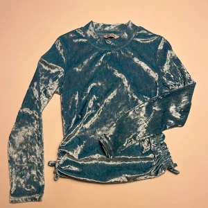 Turkos sammetströja med rynkdetaljer - Säljer en snygg turkos tröja i sammet med långa ärmar och rynkdetaljer på sidorna. Perfekt för att ge en touch av glamour till din outfit. Tröjan har en glansig finish som fångar ljuset på ett vackert sätt.