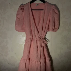 Rosa omlottklänning med knytband - Charmig rosa omlottklänning från  & other stories med korta puffärmar och knytband i midjan. Klänningen har en v-ringad design och är tillverkad i ett ribbat material som ger en fin struktur.
