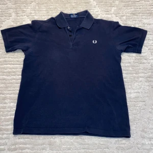 Mörkblå pikétröja från Fred Perry - Snygg mörkblå pikétröja från Fred Perry i 100% bomull. Tröjan har en klassisk krage med två knappar och det ikoniska Fred Perry-emblemet broderat på bröstet. Perfekt för en stilren och avslappnad look.