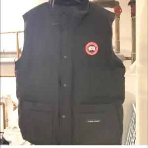 Svart dunväst från Canada Goose - Säljer en svart dunväst från Canada Goose med knappar och två praktiska fickor framtill. Västen har en klassisk design med en rund logotyp på bröstet. Perfekt för kyliga dagar.